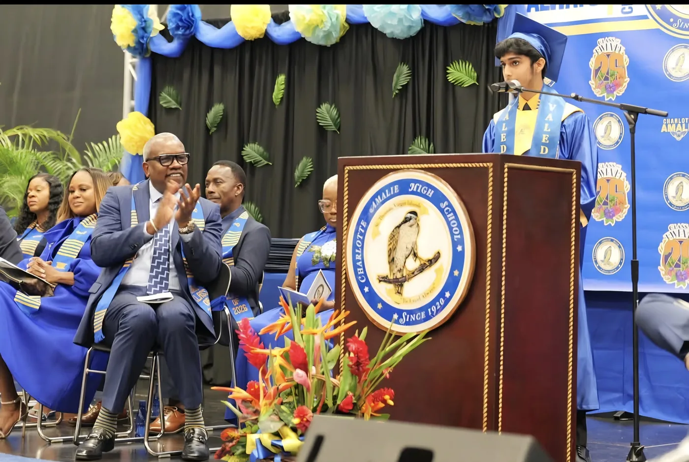 Mirza N. Baig delivering the valedictorian address at a podium bearing the Charlotte Amalie High School seal, as Governor Albert Bryan Jr. applauds from the stage.