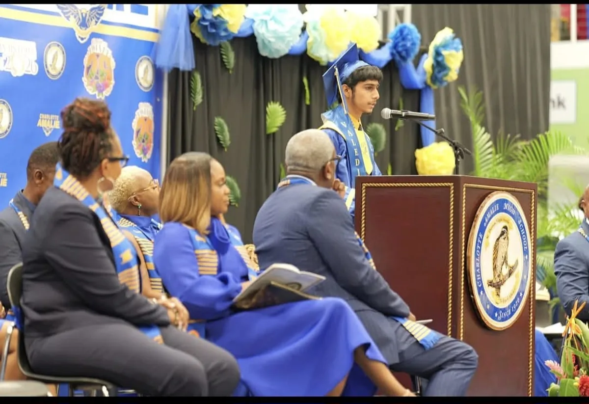 Mirza N. Baig delivering the valedictorian address at a podium bearing the Charlotte Amalie High School seal, with faculty and school officials seated on stage behind him.