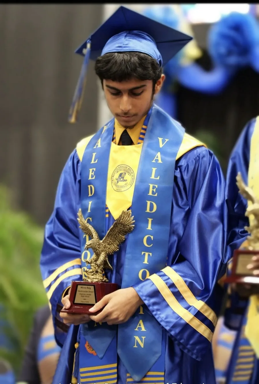 Mirza N. Baig in a blue cap and gown with Valedictorian sashes, holding the Charlotte Amalie High School valedictorian award engraved with his name and the Class of 2026.